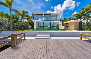 Waterfront property featuring a wood-finish dock with timber railings, an integrated in-ground swimming pool, a minimalist two-story residence with floor-to-ceiling glass windows, and a detached exterior kitchen structure