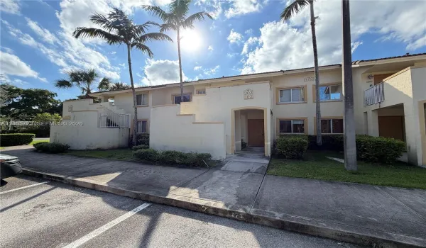 Two-story exterior featuring a light-colored stucco facade, recessed entryways, metal balcony railings, and manicured landscaping with established palm trees