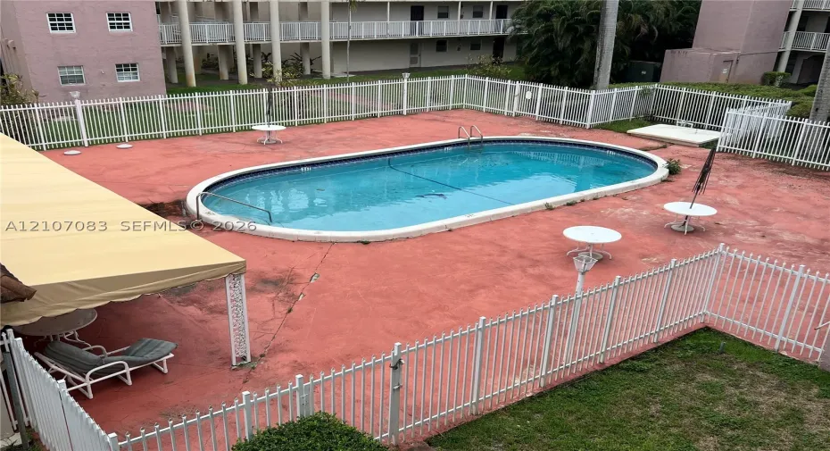 Oval-shaped swimming pool with a surrounding paved deck, metal perimeter fencing, and an attached shade structure