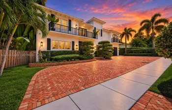 Two-story exterior featuring 2 car garage, circular drive, black balcony railings, a herringbone-patterned brick driveway, manicured landscaping, and outdoor wall mounted lighting