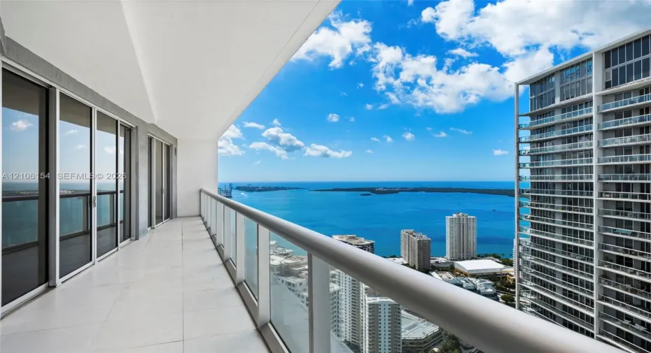 Expansive covered balcony featuring large-format tile flooring, a glass-panel railing with a minimalist handrail, and floor-to-ceiling sliding glass doors