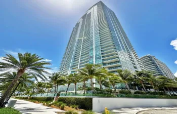 High-rise structure featuring floor-to-ceiling glass paneling, extensive wraparound balcony tiers, and contemporary glass railing systems, set against a backdrop of mature palm landscaping