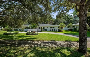 Single-story exterior featuring light-toned stucco siding, an expansive front lawn, mature canopy trees, and a concrete driveway