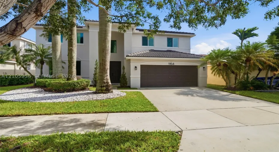 Two-story residence featuring a light-toned exterior, concrete tile roof, multi-car garage, and landscaped front grounds with mature palm trees