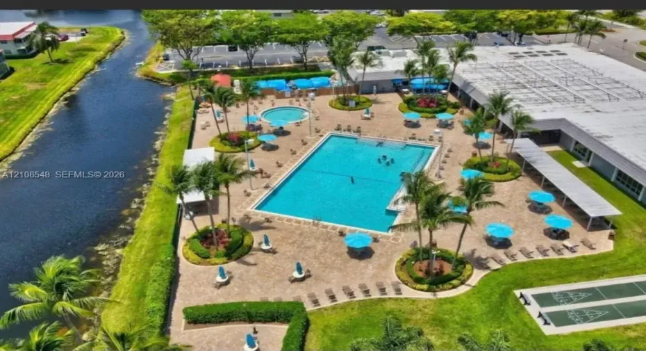 Community pool area featuring a paved deck, circular spa, mature landscaping, shuffleboard court, and canal-side positioning