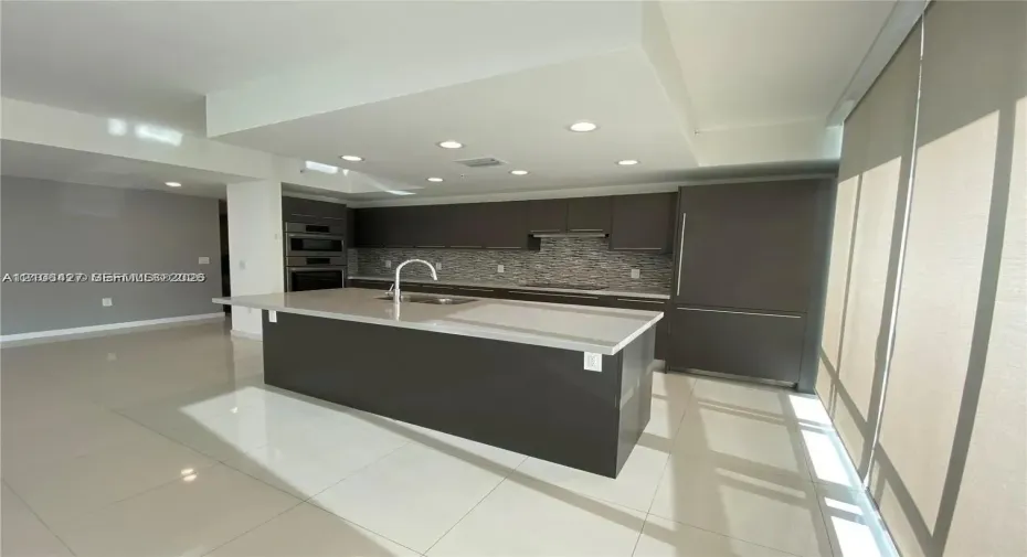 Contemporary kitchen featuring dark cabinetry, a central island with a solid-surface countertop, an integrated refrigerator, recessed ceiling lighting, and a neutral-toned backsplash