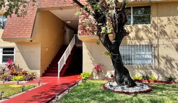 Multi-level building exterior featuring a red tile roof, red-painted concrete walkway, white-painted exterior staircase, and mature landscaping