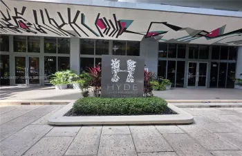 Exterior entrance featuring a structured stone-paver walkway, manicured landscaping, custom signage, and a glass-paneled facade with an artistic mural underside