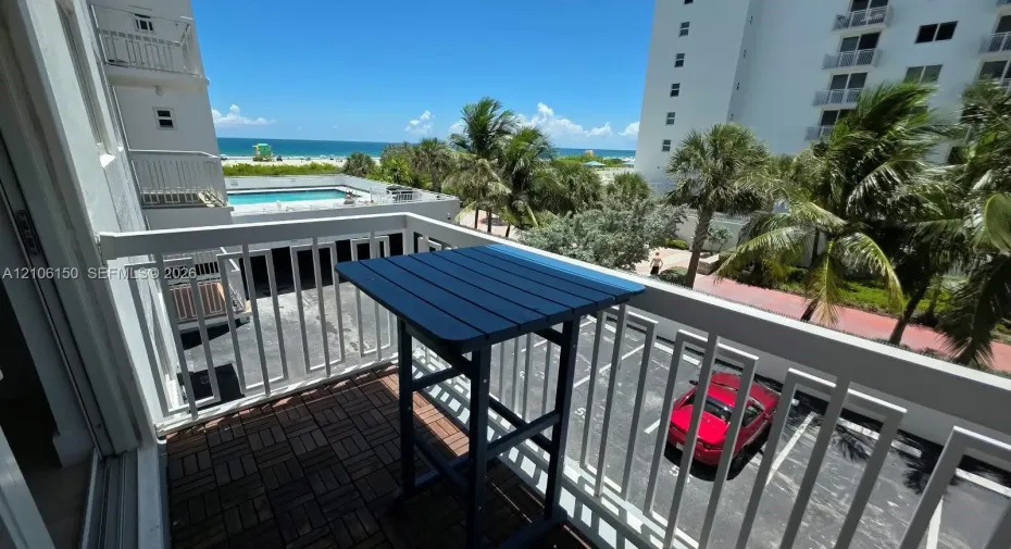 Private balcony featuring white metal railings and interlocking deck tiles, overlooking the coastline and community pool