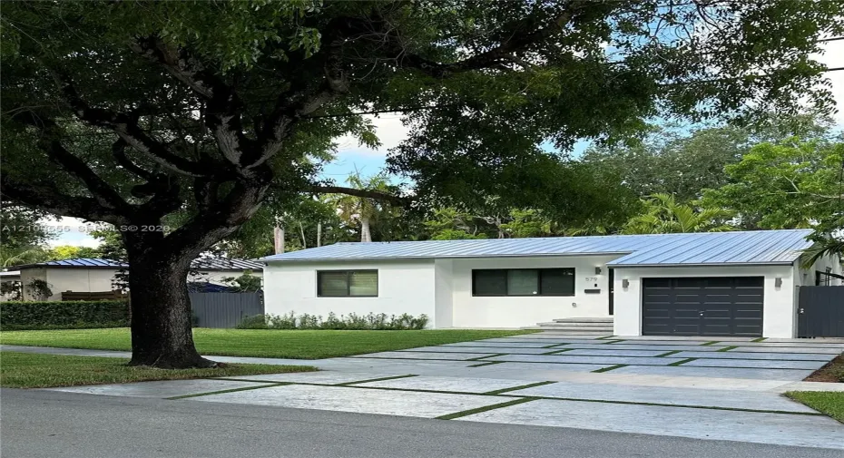Single-story exterior featuring a white stucco facade, metal roof, paver driveway with turf accents, attached garage, and mature landscaping