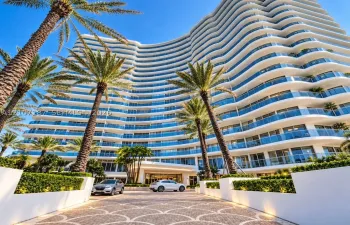 Multi-story architectural exterior featuring a curved facade, expansive balconies with glass railings, patterned stone paver motor court, and manicured landscaping with mature palms