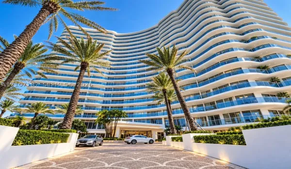Multi-story architectural exterior featuring a curved facade, expansive balconies with glass railings, patterned stone paver motor court, and manicured landscaping with mature palms