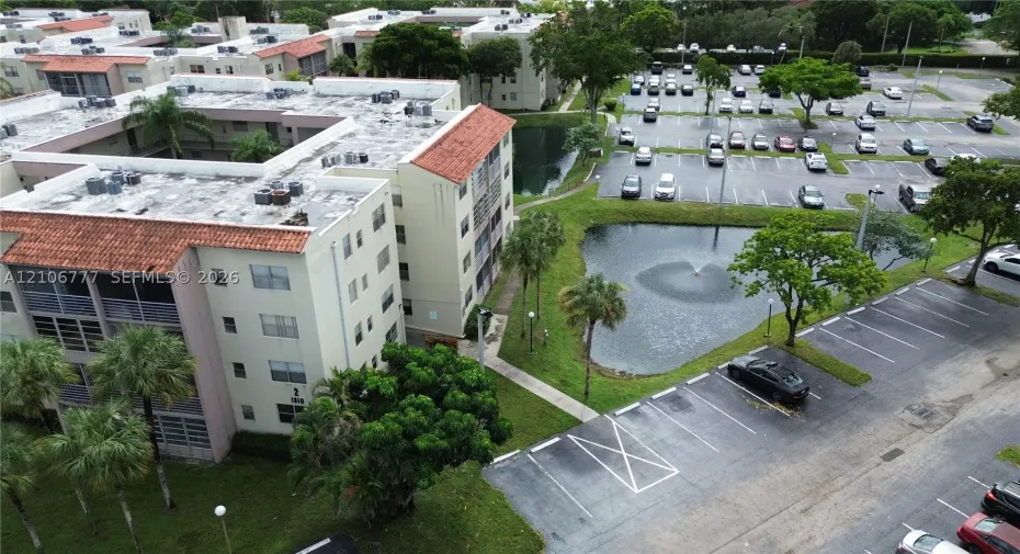 Multi-story residential building featuring a terracotta tile roof, balcony enclosures, landscaped grounds, a central fountain, and dedicated parking areas