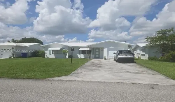 Single-story exterior featuring white stucco siding, a concrete driveway, metal window awnings, and a covered entryway