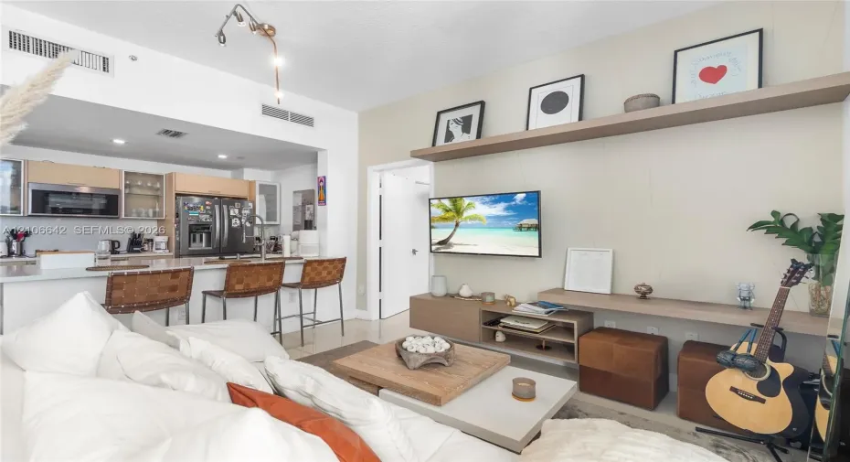 Living area featuring custom floating shelves, built-in cabinetry, and neutral-toned wall treatments, paired with an open kitchen containing integrated appliances, light-wood cabinetry, and stone-surfaced breakfast bar