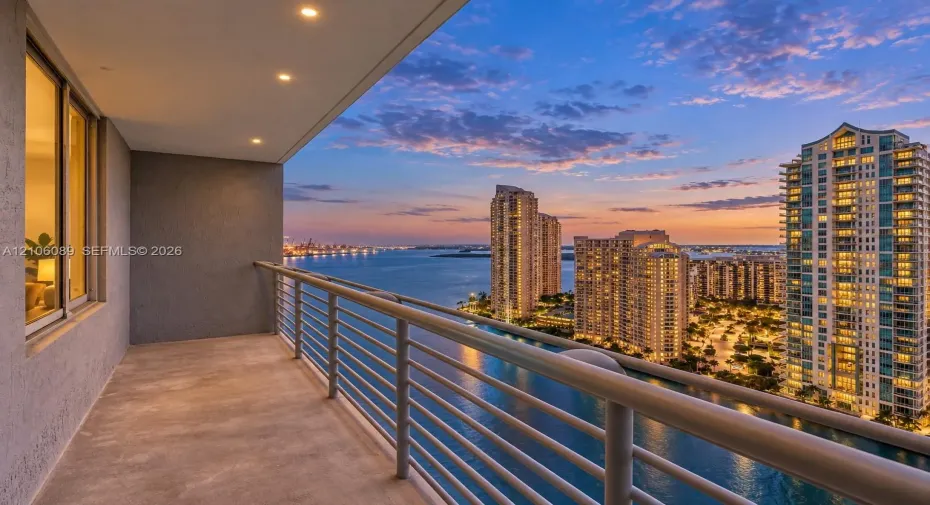 Covered balcony featuring concrete flooring, recessed lighting, and a metal horizontal-bar railing, highlighting a water and city skyline backdrop