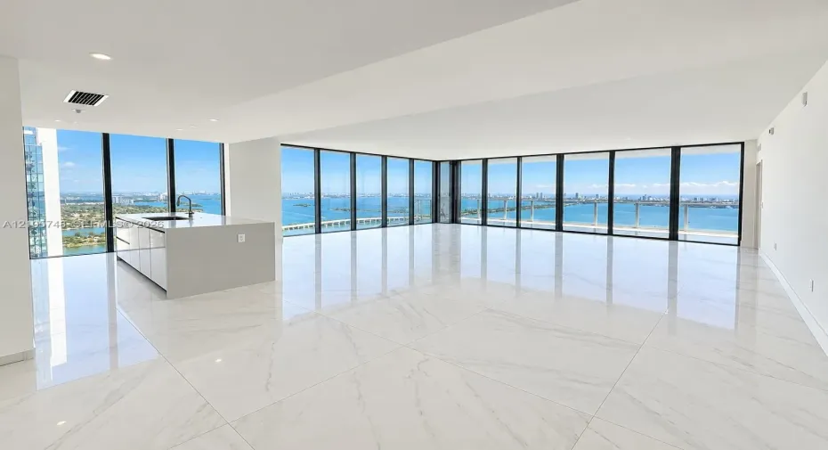 Open-concept space featuring floor-to-ceiling windows, polished stone-finish tile flooring, and a kitchen island with a neutral countertop and integrated sink