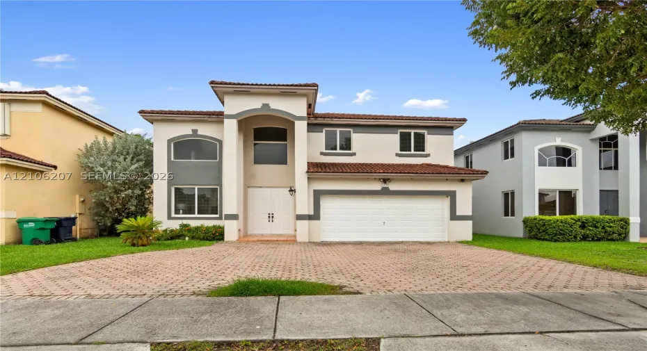 Two-story exterior featuring a clay tile roof, arched entryway, double-door main entrance, and a paved stone driveway with an attached two-car garage