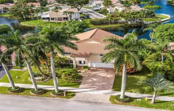 Two-story residence featuring a tile roof, brick-paver driveway, multi-car garage, mature palm landscaping, and waterfront positioning