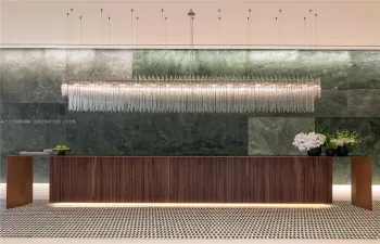 Large-scale linear chandelier with crystalline fringe, wood-veneer floating desk with base accent lighting, and full-height green-toned stone feature wall