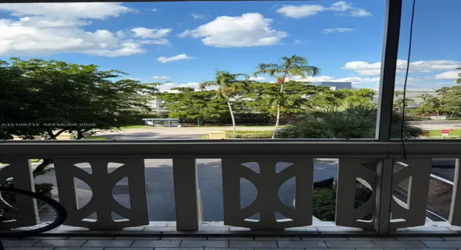 Screened balcony featuring decorative white geometric railing and tiled flooring, overlooking mature palm trees and green space