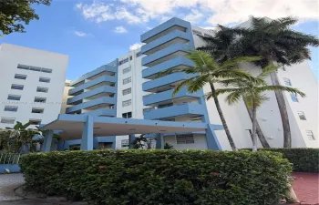 Multi-story residential building exterior featuring stacked private balconies, a structural covered entryway, and mature palm trees