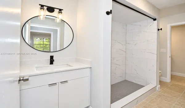 White vanity with stone-composite countertop, undermount sink, matte black faucet, round mirror with black frame, and walk-in shower featuring large-format patterned wall tile and dark hexagonal floor tile