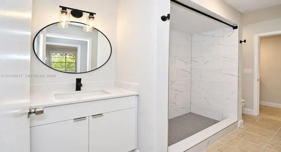 White vanity with stone-composite countertop, undermount sink, matte black faucet, round mirror with black frame, and walk-in shower featuring large-format patterned wall tile and dark hexagonal floor tile