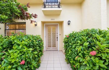 Entryway featuring a decorative metal-grille front door, wrought iron balcony railing, stucco exterior, ceramic tile walkway, and wall-mounted exterior lighting