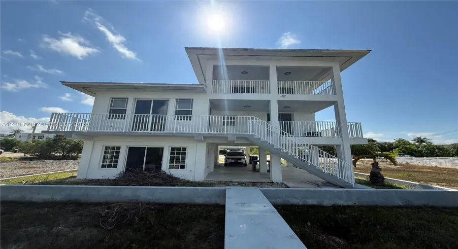 Multi-level stilt-style residence featuring a white stucco exterior, multiple covered balconies with white metal railings, a concrete ground-level carport, and a concrete walkway