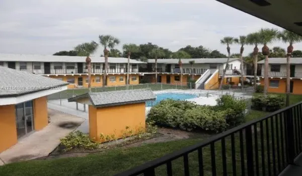 Swimming pool with metal perimeter fencing, multi-level building exterior with walkway balconies, and mature palm trees