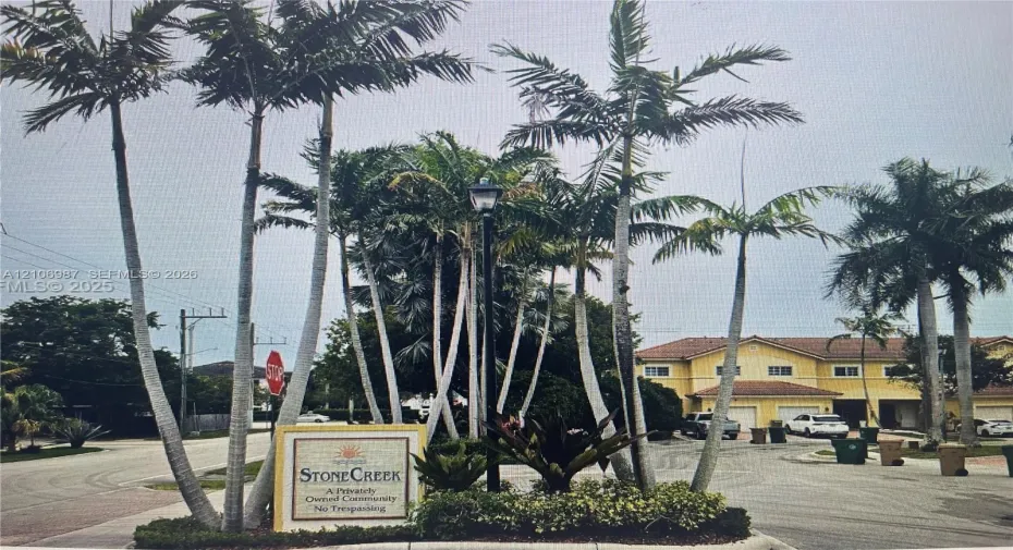 Stone Creek community entrance featuring mature palm trees, professional landscaping, a monument sign, and asphalt paving
