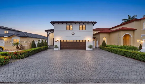 Stucco exterior residence featuring a herringbone-pattern paver driveway, dark-tinted sectional garage door, clay tile roofing, and arched entryway