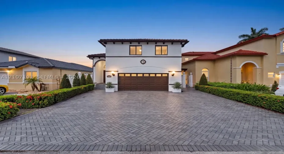 Stucco exterior residence featuring a herringbone-pattern paver driveway, dark-tinted sectional garage door, clay tile roofing, and arched entryway