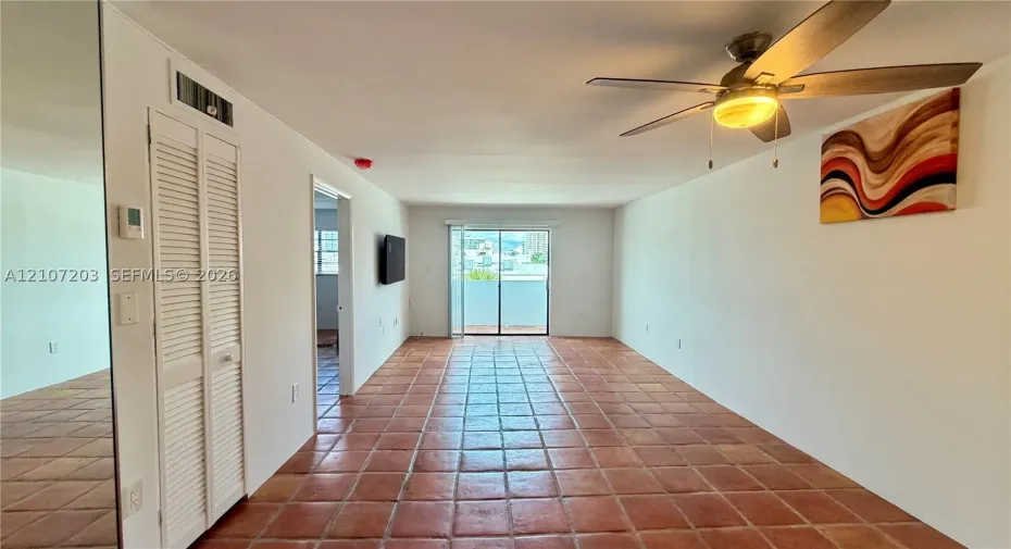 Large open-concept room featuring terra cotta tile flooring, floor-to-ceiling mirror paneling, a ceiling fan with integrated lighting, and sliding glass doors