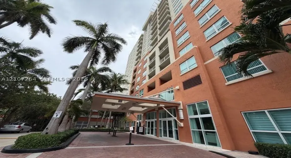 High-rise exterior featuring a structured porte-cochère, textured masonry facade, multi-story glass fenestration, and landscaped palm trees along a paver stone arrival court
