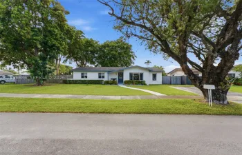 Single-story residence featuring light-colored exterior siding, a wide front lawn, mature shade trees, a concrete walkway, and private perimeter fencing