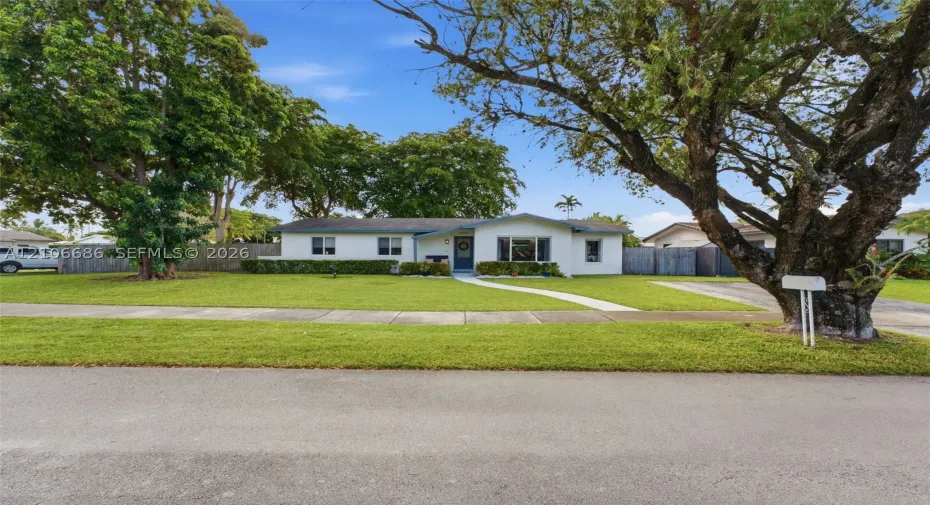 Single-story residence featuring light-colored exterior siding, a wide front lawn, mature shade trees, a concrete walkway, and private perimeter fencing