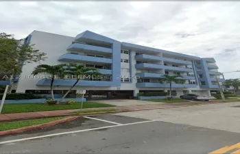 Multi-story residential building featuring blue-accented exterior, private balconies, covered ground-level parking, and landscaped grounds