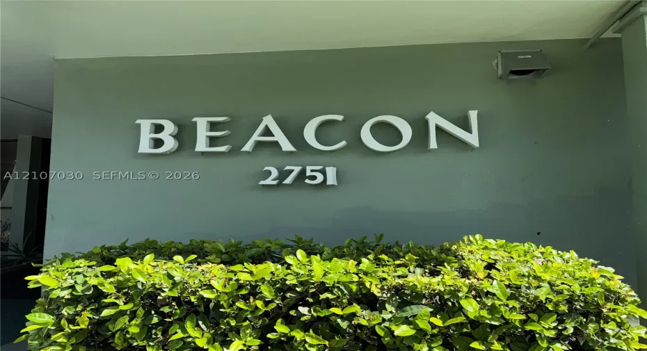 Dimensional building signage featuring raised lettering and address numbers set against a solid-tone exterior wall, accented by a manicured shrub hedge