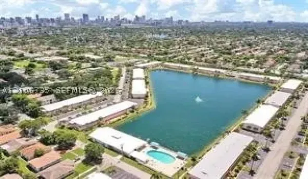 Aerial perspective featuring a multi-building residential complex surrounding a central lake with a fountain, including a swimming pool and paved parking areas