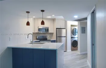 Kitchen featuring an island with a stone-finish countertop, stainless steel appliances, neutral cabinetry, stacked laundry unit, and wood-finish flooring