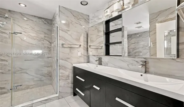Dual-sink vanity with dark cabinetry and metallic hardware, large wall-to-wall mirror, glass-enclosed shower, and full-height wall tile