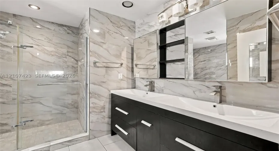 Dual-sink vanity with dark cabinetry and metallic hardware, large wall-to-wall mirror, glass-enclosed shower, and full-height wall tile