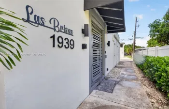 Exterior facade featuring smooth stucco finish, custom metal address signage, wall-mounted cylindrical sconces, dark metal awning, and louvered entry door