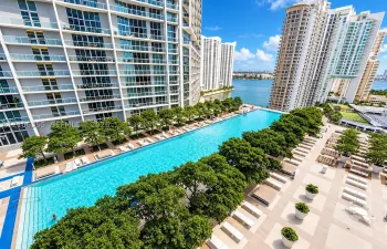 Oversized lap pool with tiered entry steps, extensive paved pool deck, and mature canopy trees, set against a waterfront backdrop
