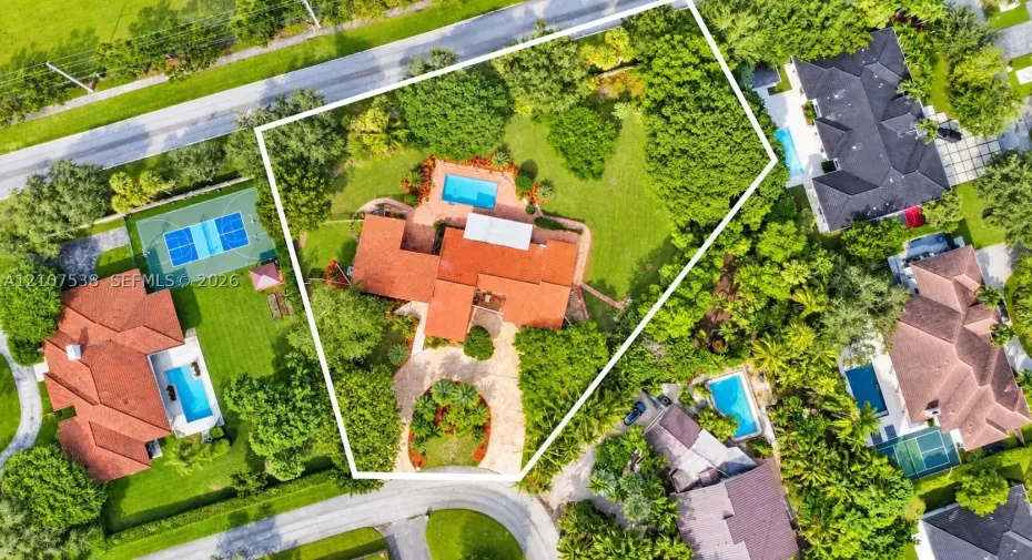 Aerial perspective of a sprawling property featuring a multi-level clay tile roof, a circular paver driveway with a central landscape island, and an in-ground swimming pool