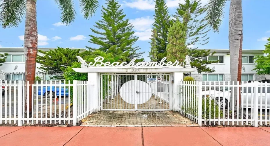 Gated entrance featuring white decorative metal fencing, stone-paver walkway, and mature palms