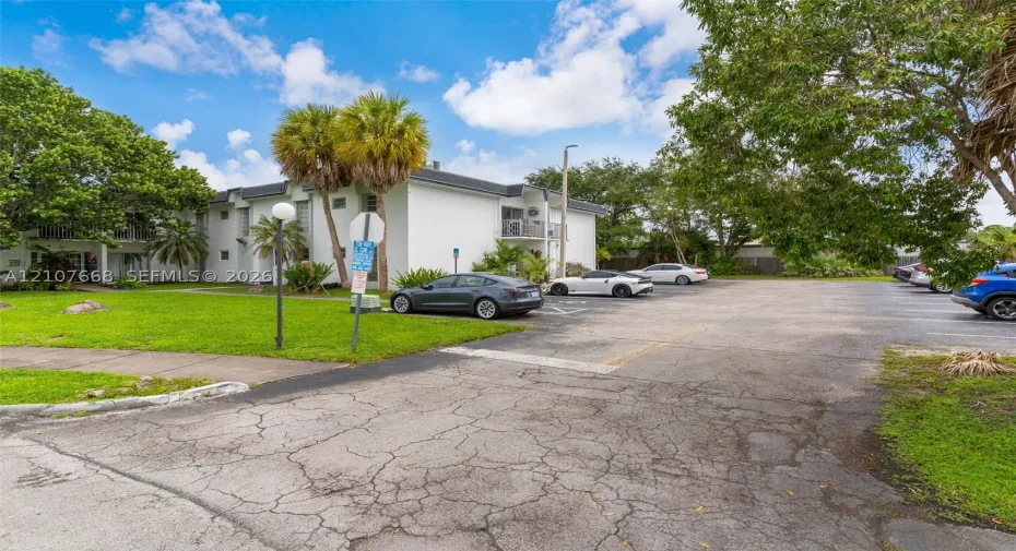 Multi-story building exterior featuring smooth white siding, paved surface parking, and mature palm and shade trees