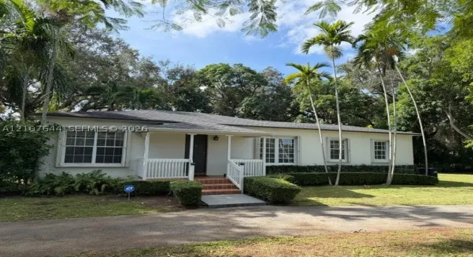 Single-story residence featuring a covered front porch with a white balustrade, brick entry steps, multi-pane windows, and manicured landscaping with mature palm trees